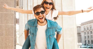 Smiling beautiful girl and her handsome boyfriend in casual summer clothes. Man carrying his girlfriend on the back and she raising her hands.Happy cheerful family having fun on the street background