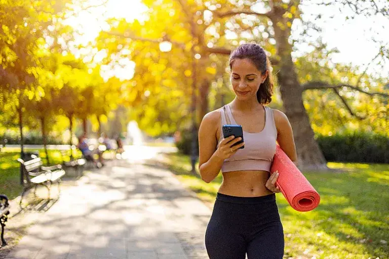 Фит девојка во спортска облека спремна за јога на утринска прошетка со мобилен телефон во рака и насмевка на лицето
