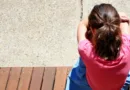 Деовјче во црвена маица како седи осамено на клупа, сликано (позади) во грб - не се гледа лице